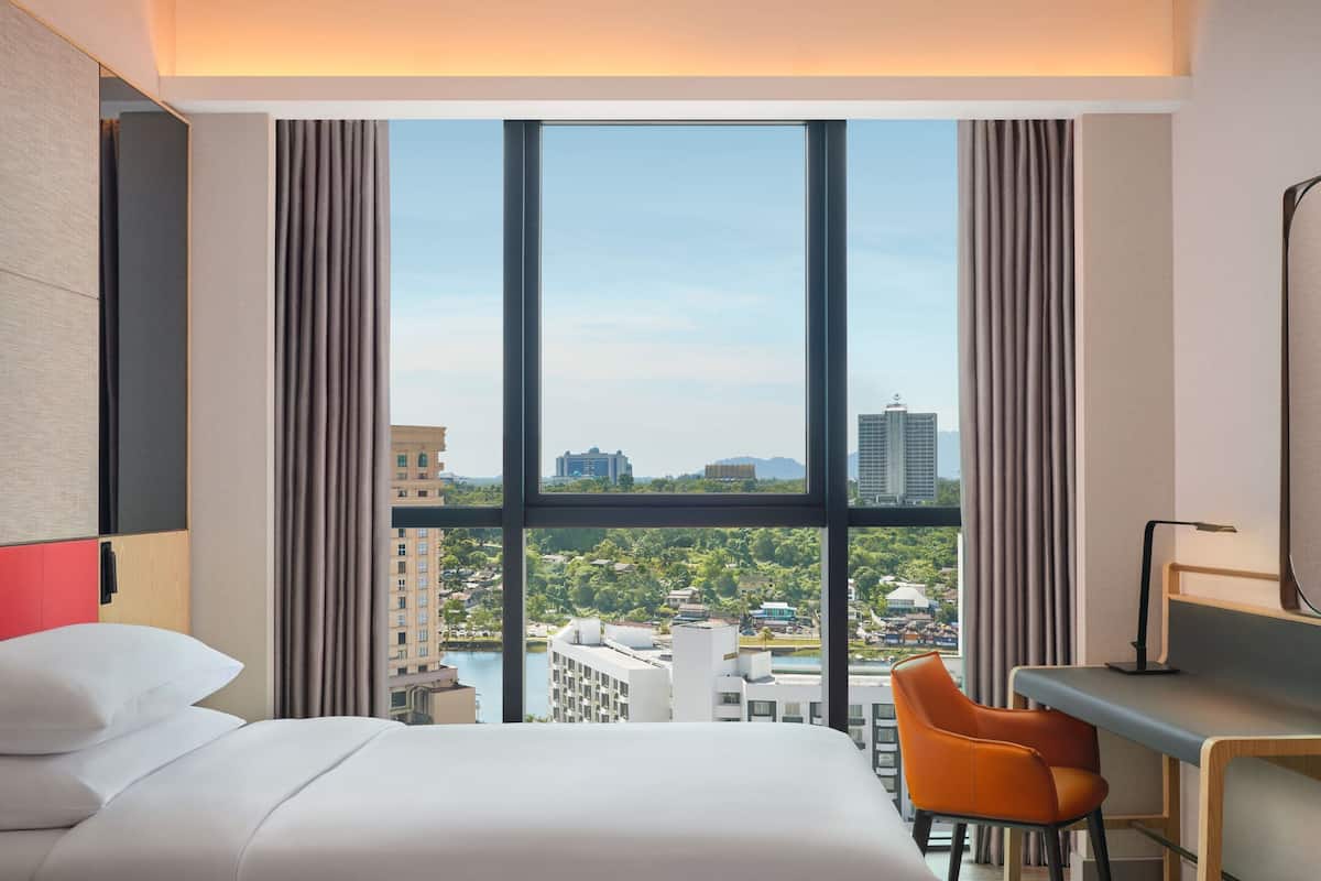 A hotel room with a large window, a bed, an orange chair, and a desk with a lamp.