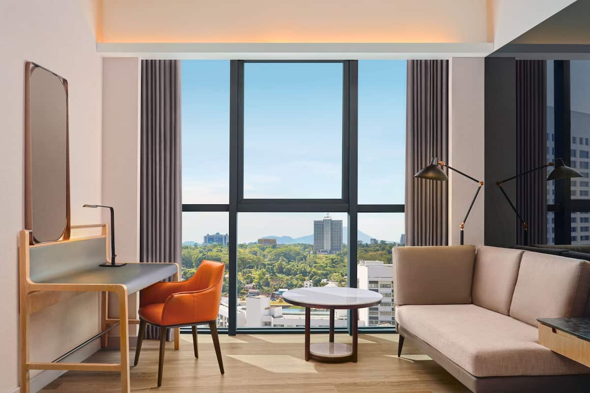A modern hotel room with a large window, a sofa, a desk, and a chair.