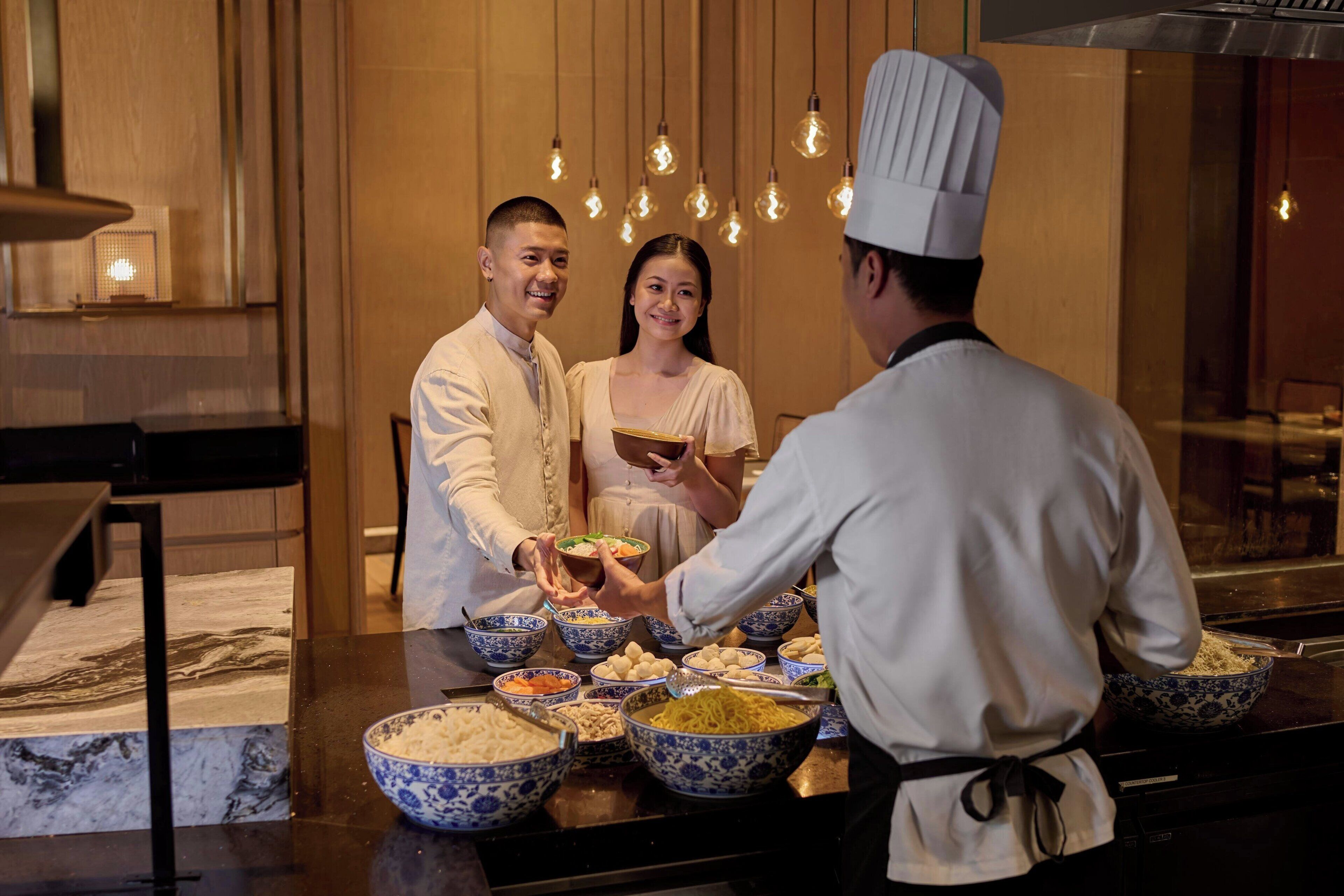A chef serving food to guests in a restaurant with a marble countertop and hanging light bulbs.