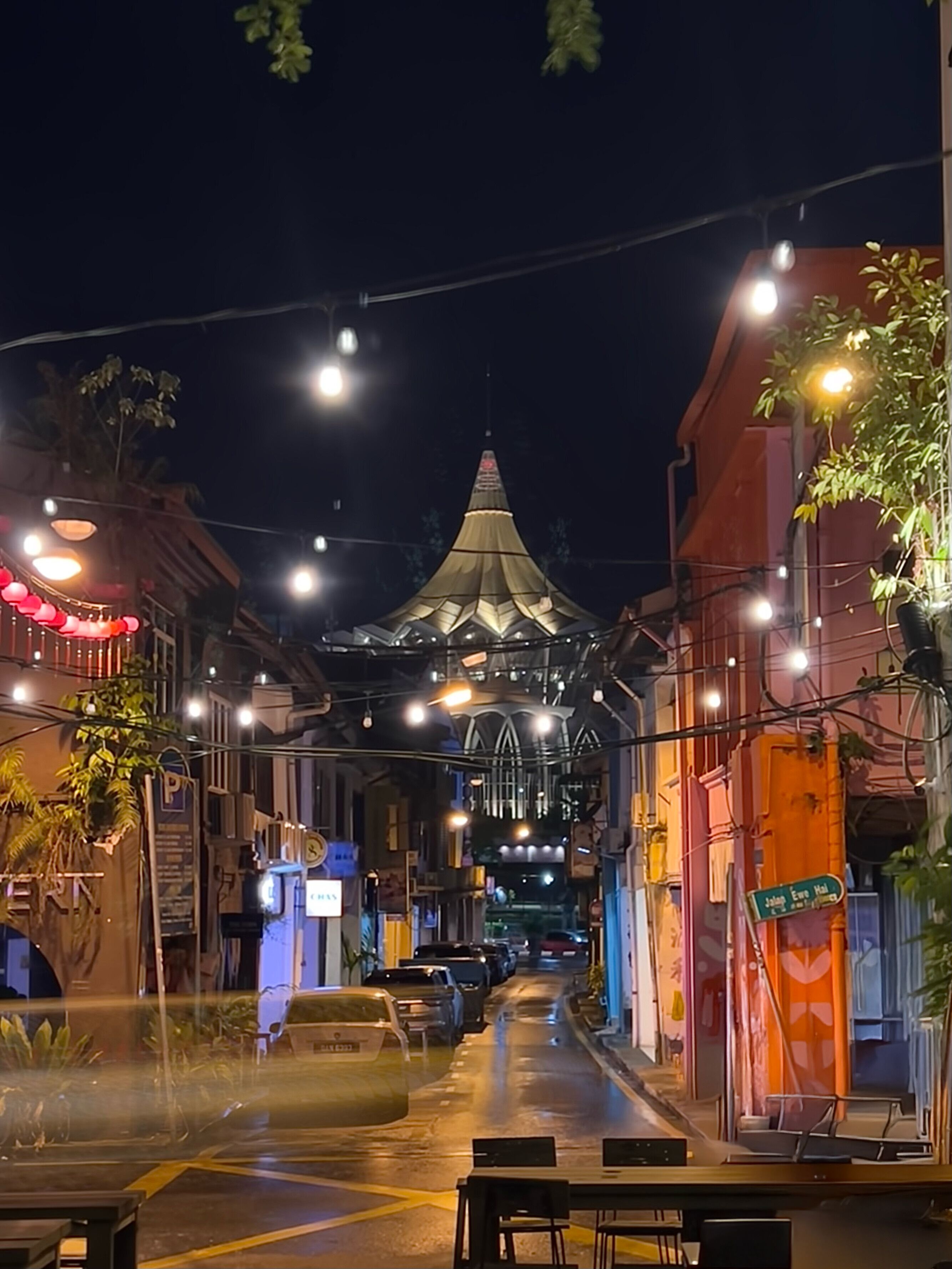 A street at night with a prominent building featuring a pointed roof and illuminated by string lights.