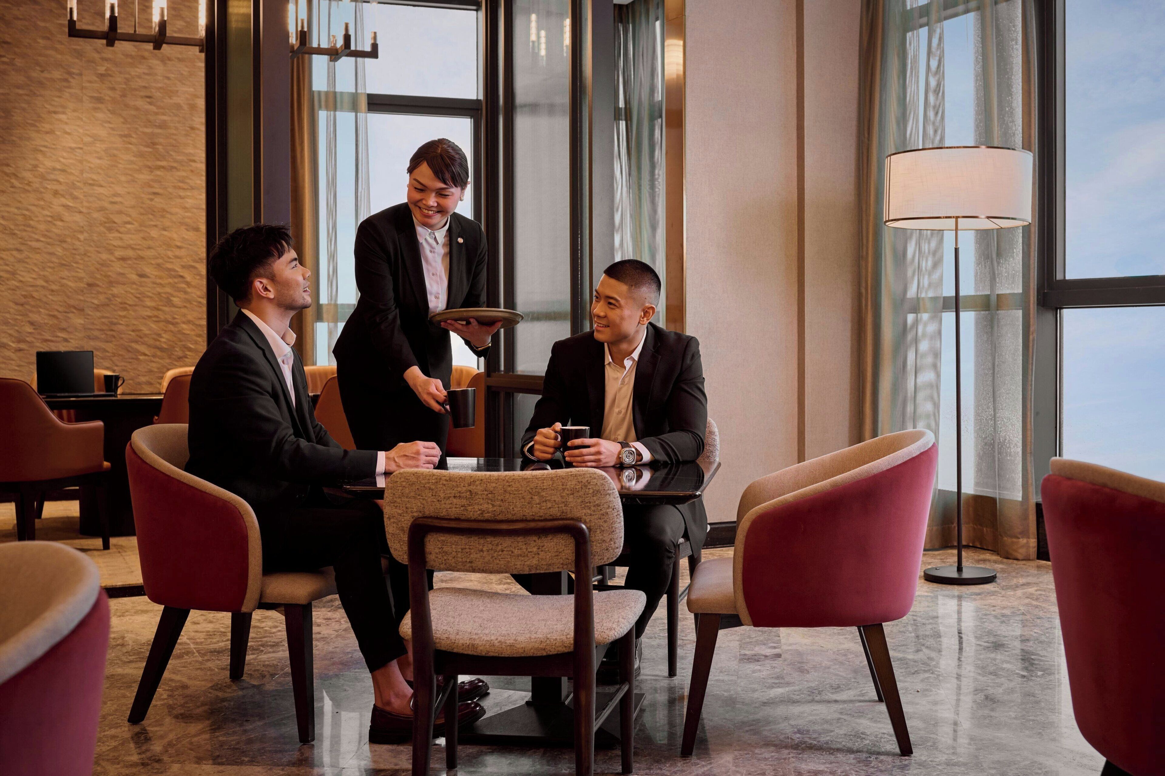 A group of three people seated around a table in a modern hotel lobby with large windows and a floor lamp.
