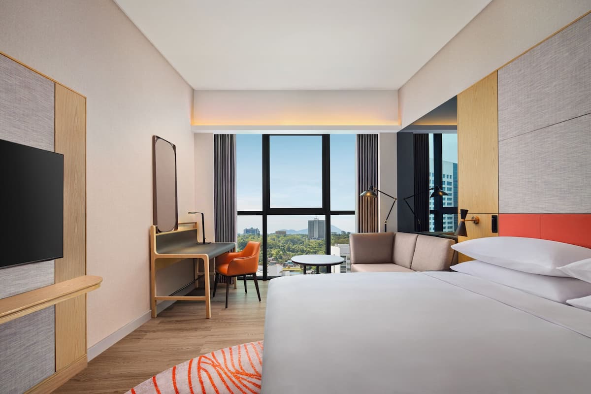 A modern hotel room with a large bed, a desk, a chair, a TV, and a view of the city through large windows.