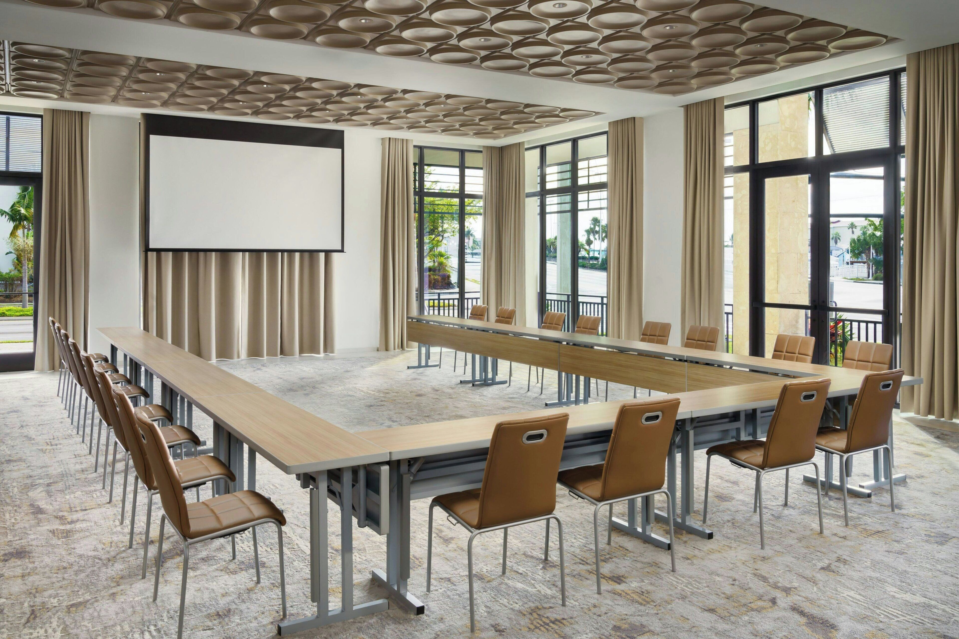 A conference room with a long table, chairs, a projector screen, and large windows with curtains.