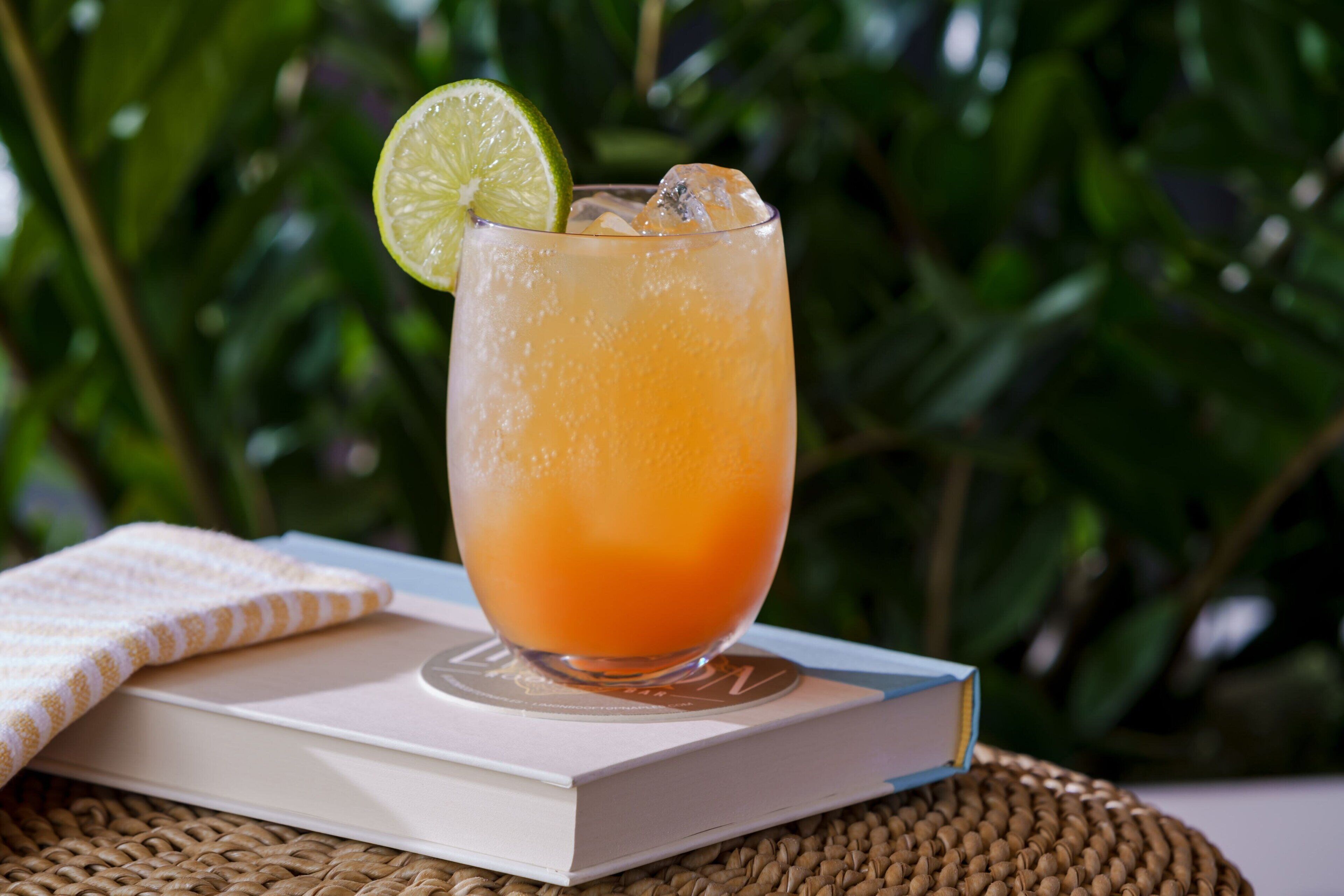 A glass of orange beverage with a lime slice on a coaster, placed on a book with a woven mat underneath.