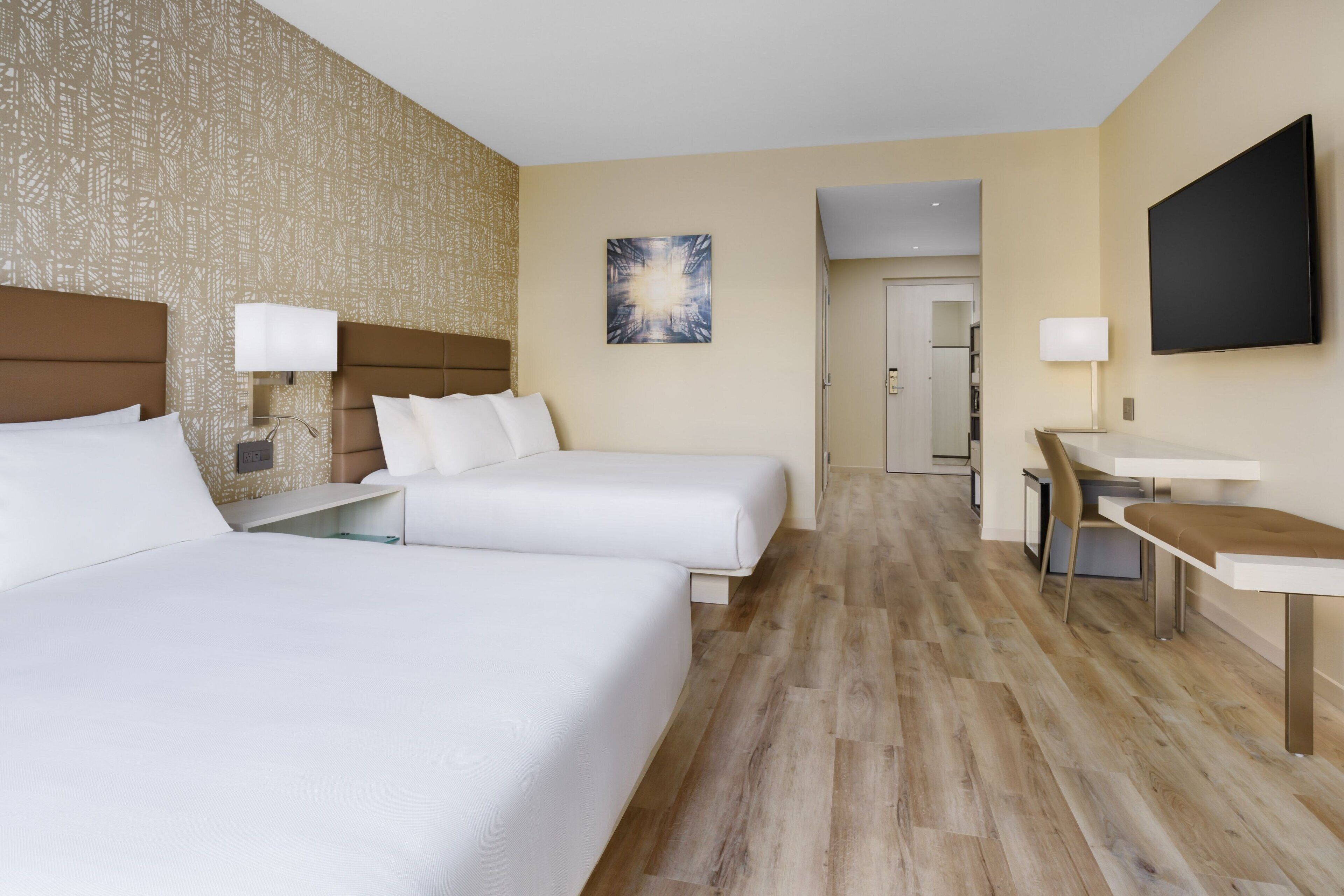 Quarto de hotel com duas camas, uma escrivaninha, uma cadeira, uma televisão e uma porta para o banheiro.