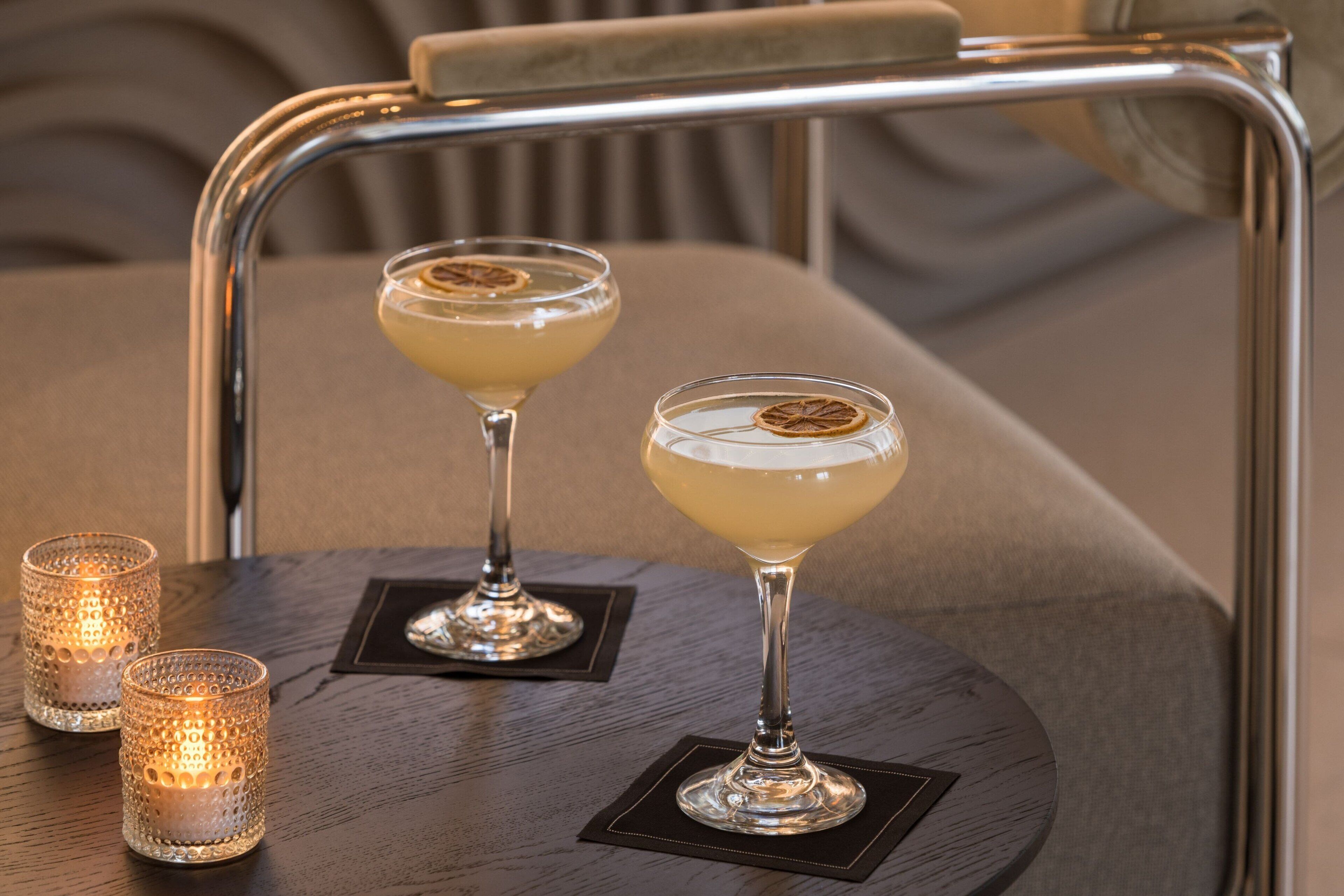 Deux verres à cocktail sont posés sur une table ronde, accompagnés d’un plateau en métal et de deux bougies allumées.