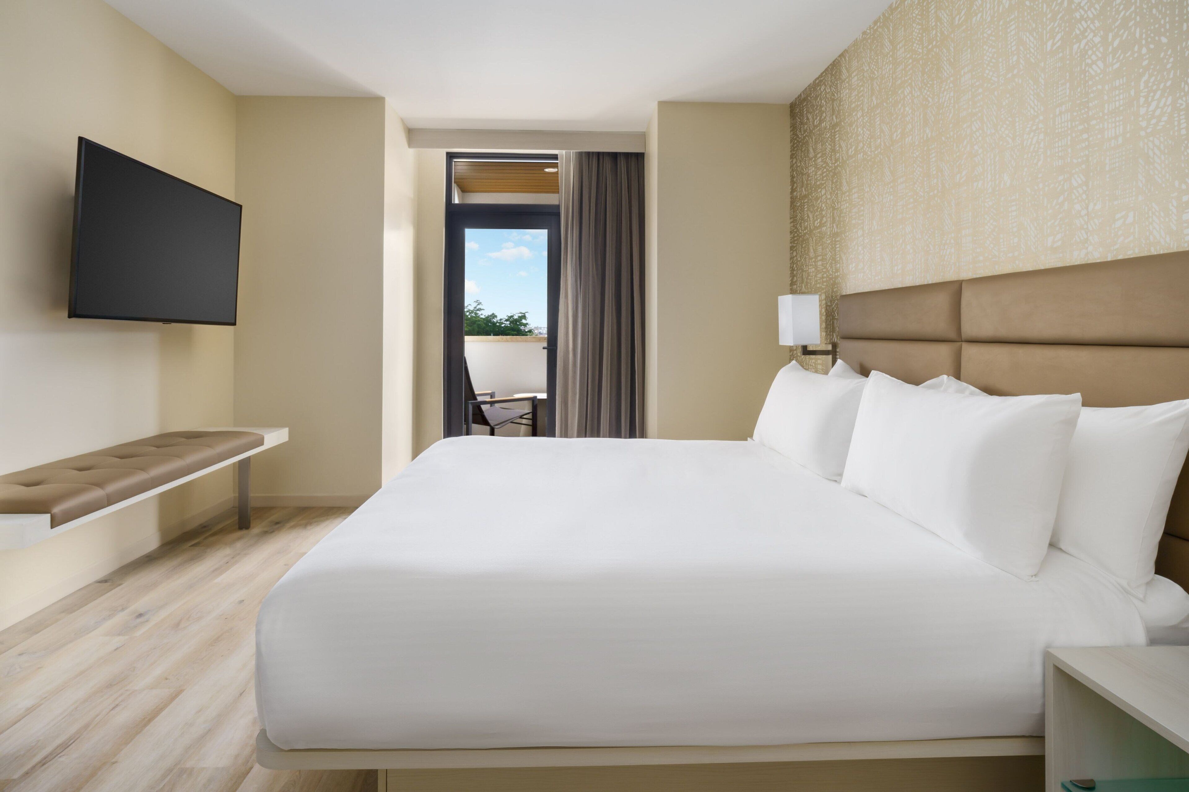 A hotel room with a large bed, a flat-screen TV mounted on the wall, a bench, and a view of the outdoors.