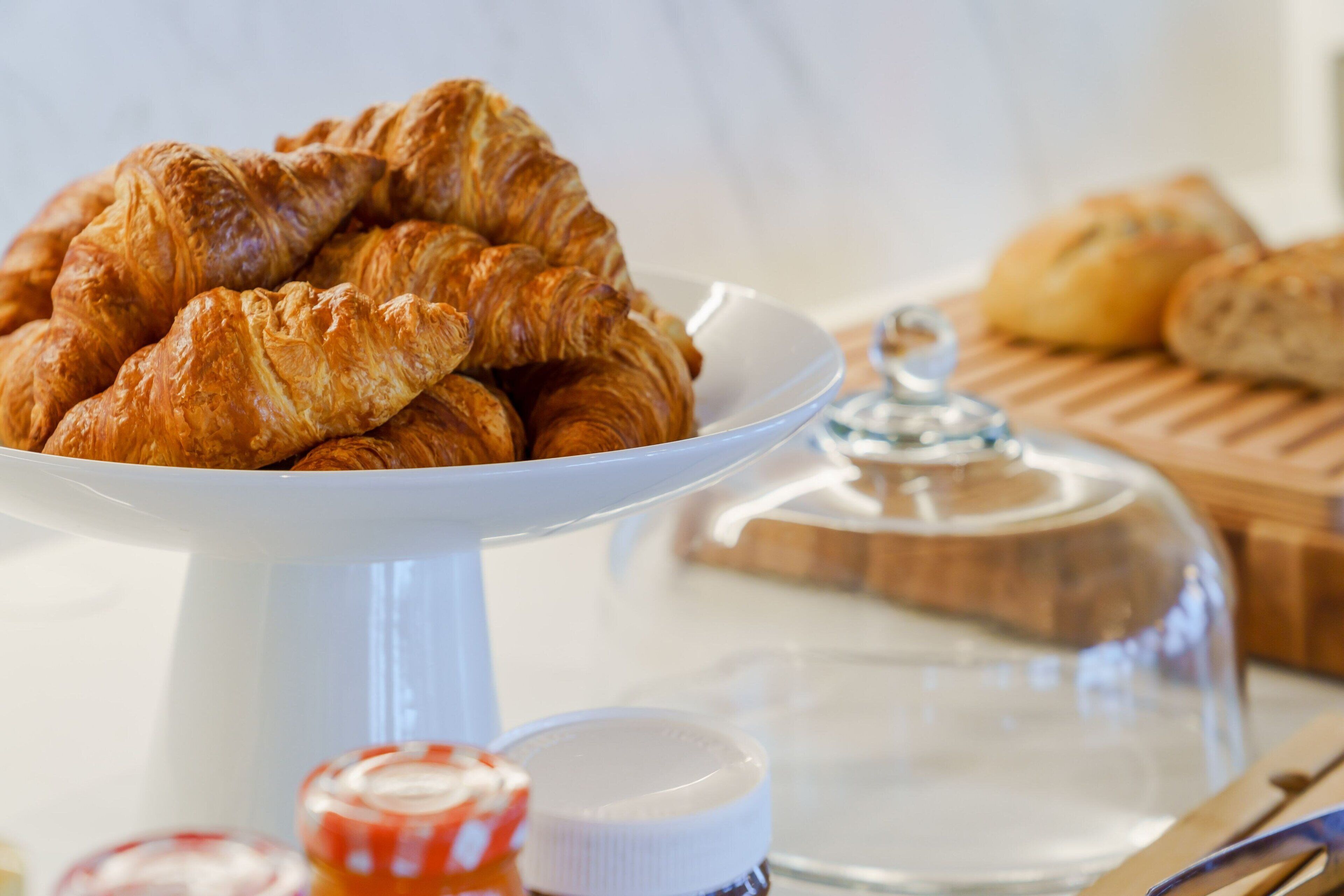 Une assiette en céramique blanche avec des croissants, un bocal en verre et une planche à découper en bois avec un couvercle en verre à l’arrière-plan.