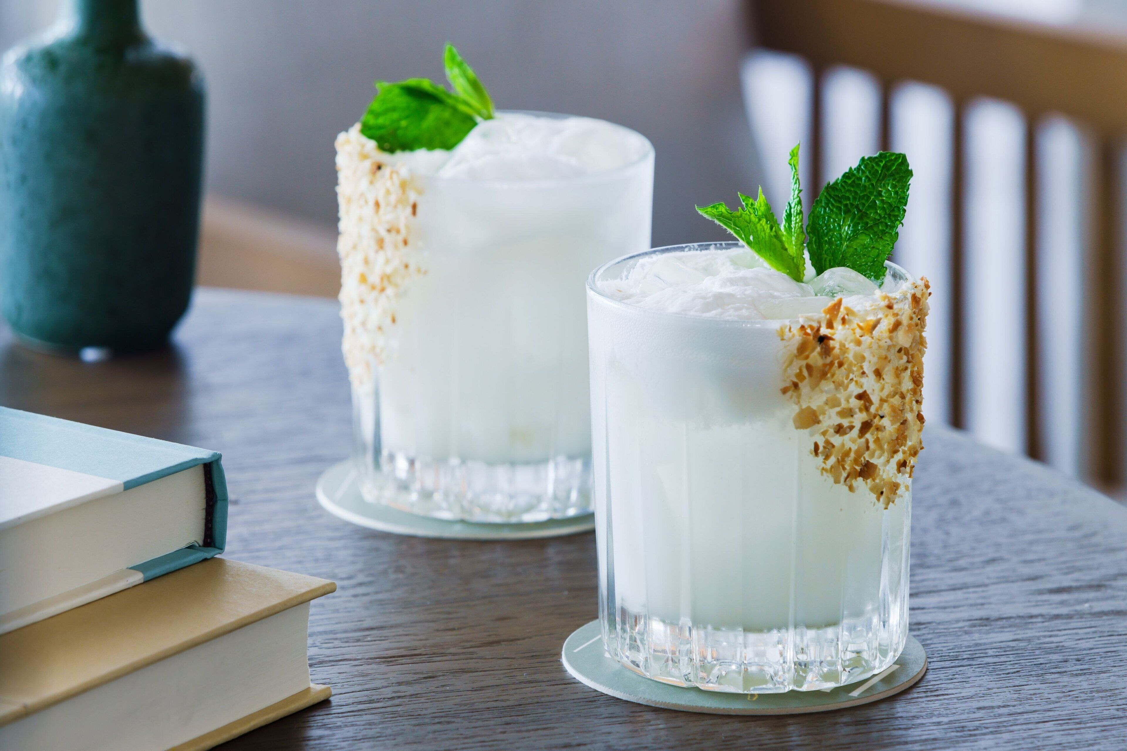 Twee glazen met een romig drankje, versierd met muntblaadjes en geroosterde kokosvlokken, op een houten tafel met een boek en een vaas op de achtergrond.