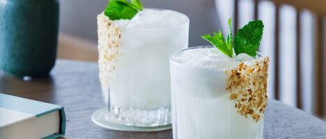 Two glasses of a creamy beverage with mint leaves, garnished with toasted coconut flakes, on a wooden table with a book and a vase in the background.