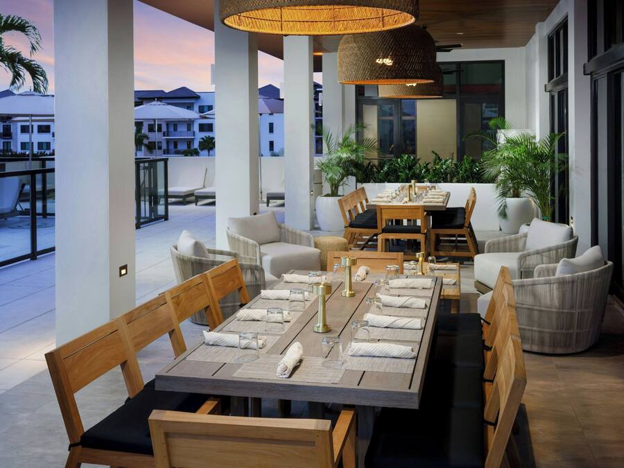A dining area with a long table set for six, surrounded by cushioned chairs, with a view of the cityscape in the background.