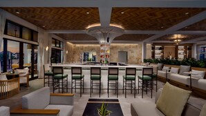 Un hall d’hôtel moderne doté d’un bar central, de différents espaces aménagés pour s’asseoir et offrant une vue sur le paysage urbain.