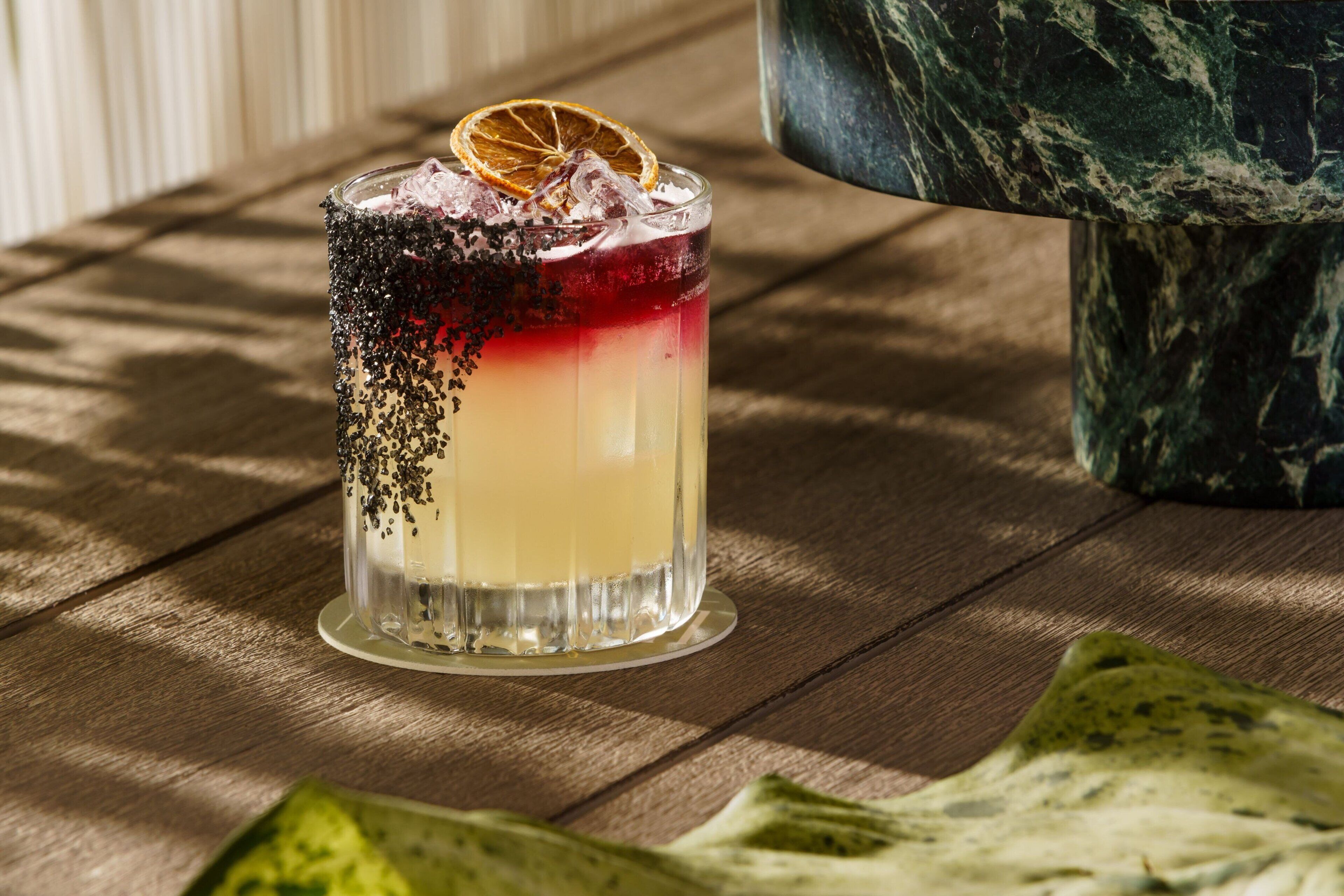 A clear glass with a layered drink, ice, and a dried orange slice garnish on a wooden surface.