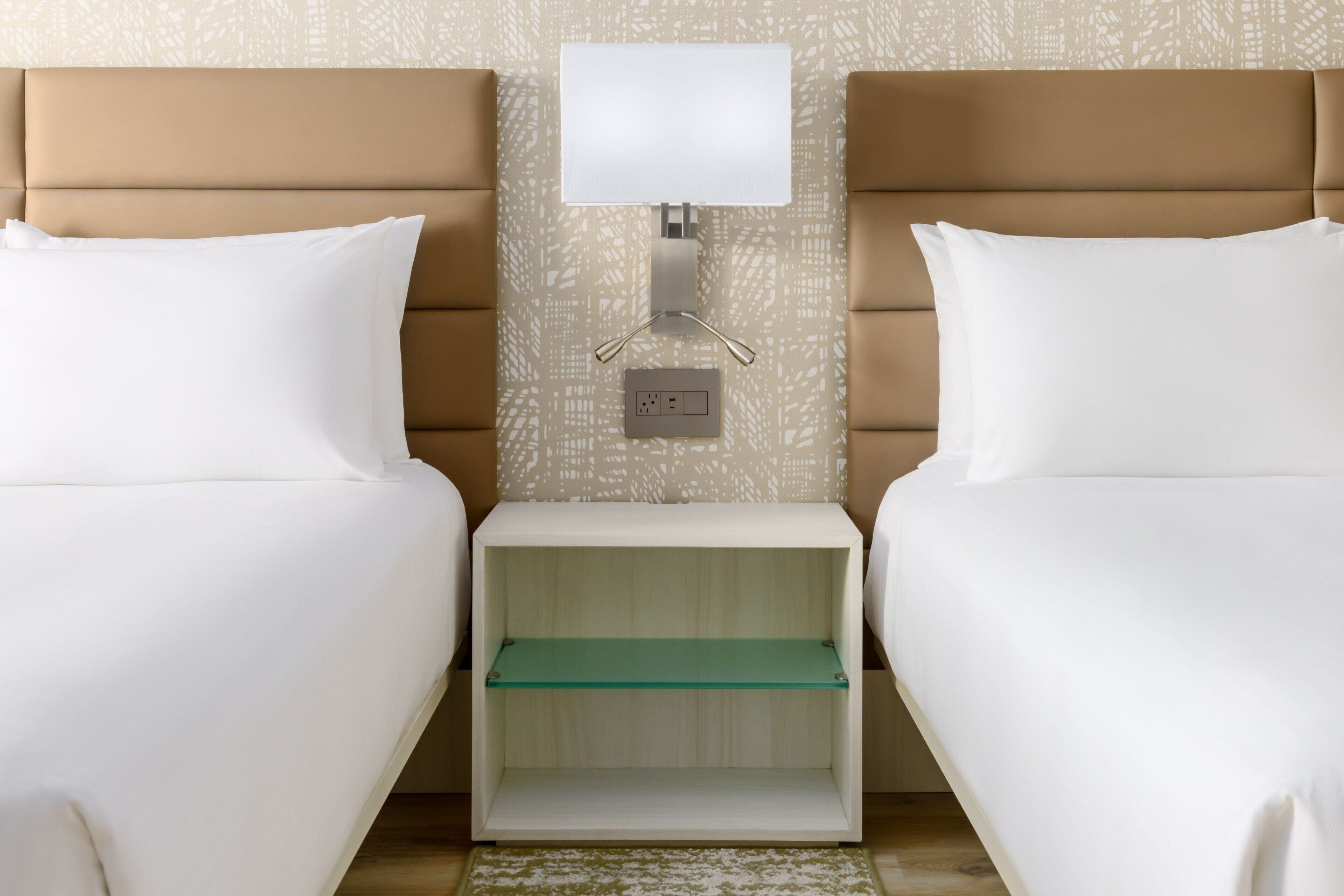 Una habitación de hotel con dos camas, una mesita de noche con lámpara y una pequeña estantería.