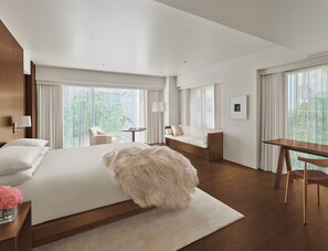 Une chambre d’hôtel moderne dotée d’un grand lit, d’un coin salon avec un canapé et des fauteuils, d’une table à manger et de grandes fenêtres offrant une vue sur la verdure.
