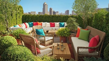 An outdoor seating area with wicker furniture and cushions, surrounded by greenery and a city skyline in the background.