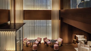 Un vestíbulo de hotel moderno con piso de madera, una zona de estar con mesas y sillas redondas, y ventanas amplias con cortinas translúcidas.