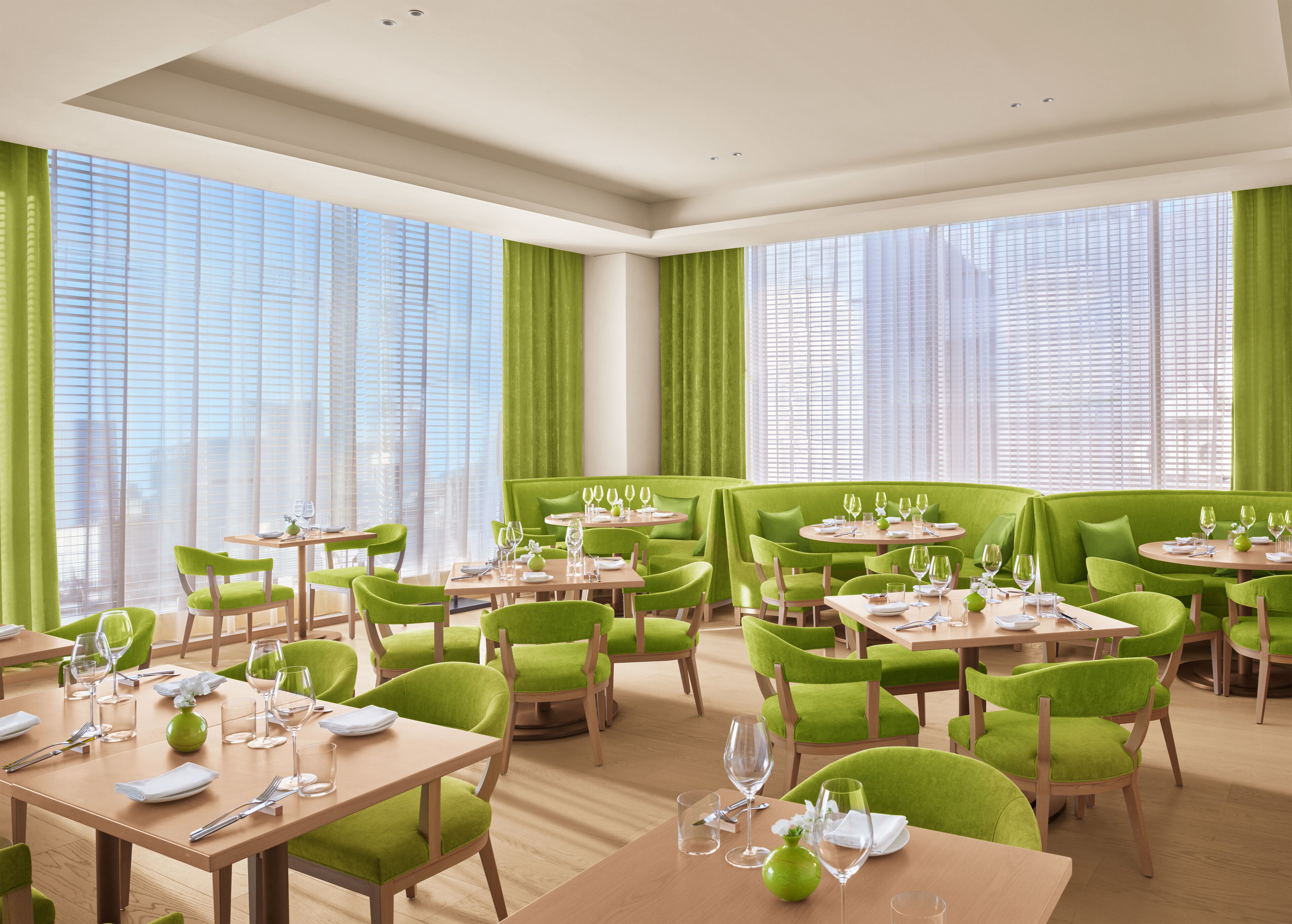 Un espace de restauration moderne avec des chaises et des tables à revêtement vert, de grandes fenêtres ornées de rideaux légers, et un design épuré et minimaliste.
