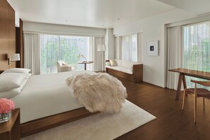 Une chambre d’hôtel moderne dotée d’un grand lit, d’un coin salon avec une table et des chaises, et de grandes fenêtres offrant une vue sur la verdure.