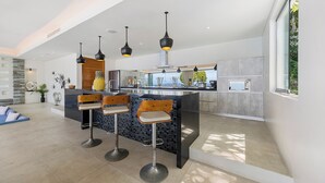 A modern kitchen with a central island, bar stools, pendant lighting, and a view of the outdoors. - Villa Spice (Koh Samui)