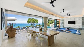 A modern living room with a large glass door leading to a balcony with a view of the ocean. There is a wooden dining table with chairs, a sofa, and a TV mounted on the wall.