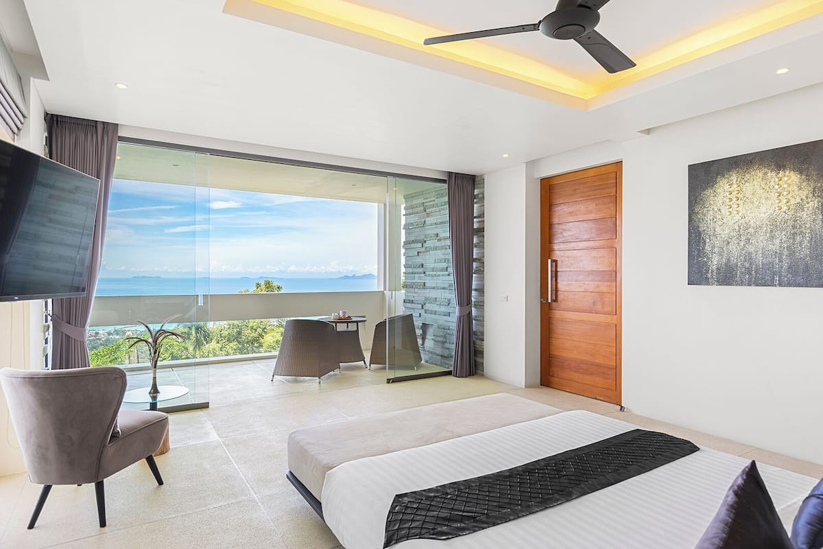 A modern bedroom with a large bed, a TV, a balcony with a view, and a wooden door.