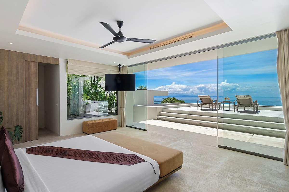 A modern bedroom with a large bed, a TV, and a balcony with a view of the ocean.