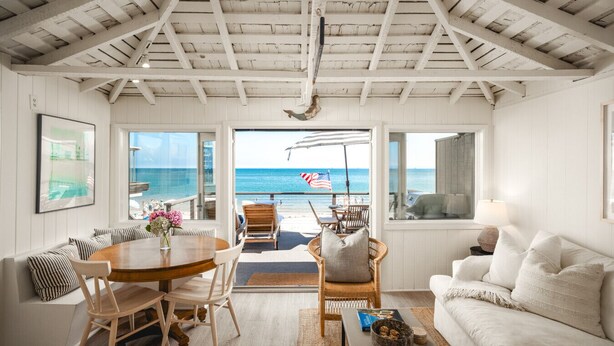 A spacious living area with a view of the ocean, a dining table with chairs, a sofa, and a coffee table with books. - Oceanfront Cottage on the Sand at Miramar Beach (Santa Barbara)