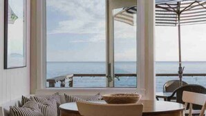 A room with a large window overlooking the sea, a wooden table with chairs, and a sofa with striped cushions. - Oceanfront Cottage on the Sand at Miramar Beach (Santa Barbara)