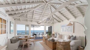 A spacious living area with a high ceiling, a dining table, and a sofa. A large window offers a view of the ocean. - Oceanfront Cottage on the Sand at Miramar Beach (Santa Barbara)