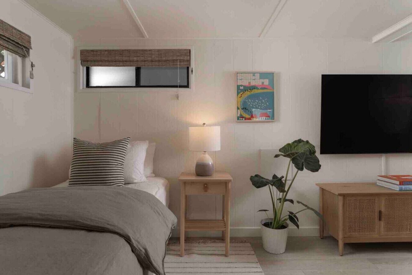 A bedroom with a bed, bedside table, lamp, potted plant, and a TV on the wall.