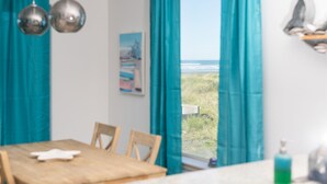 A room with a wooden table, chairs, teal curtains, a view of the beach, and a painting on the wall. - Dog-Friendly Beach Escape - 20% off March stays (Moclips)