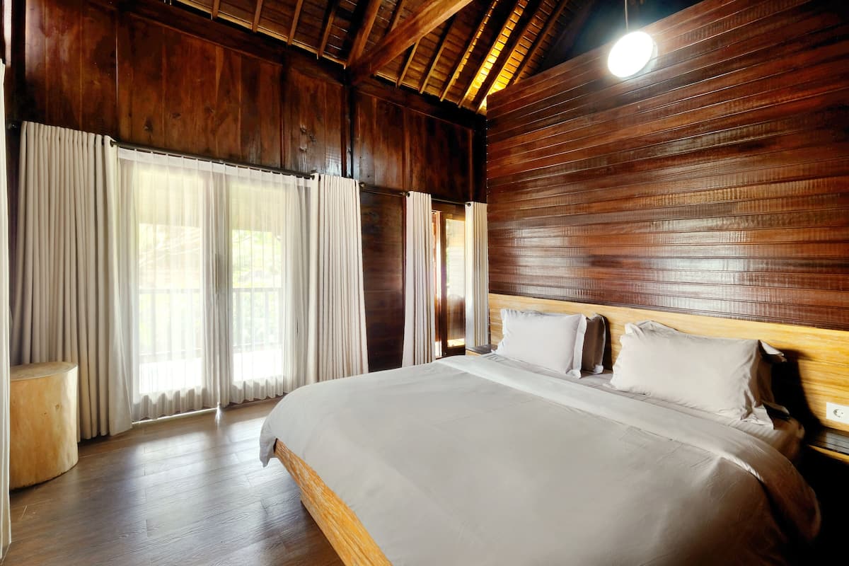 A large bed with a wooden headboard, a wooden floor, and a large window with curtains.