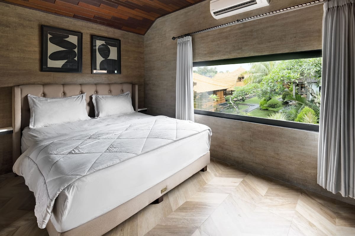 A bedroom with a large bed, a view of a garden through a window, and a ceiling fan.