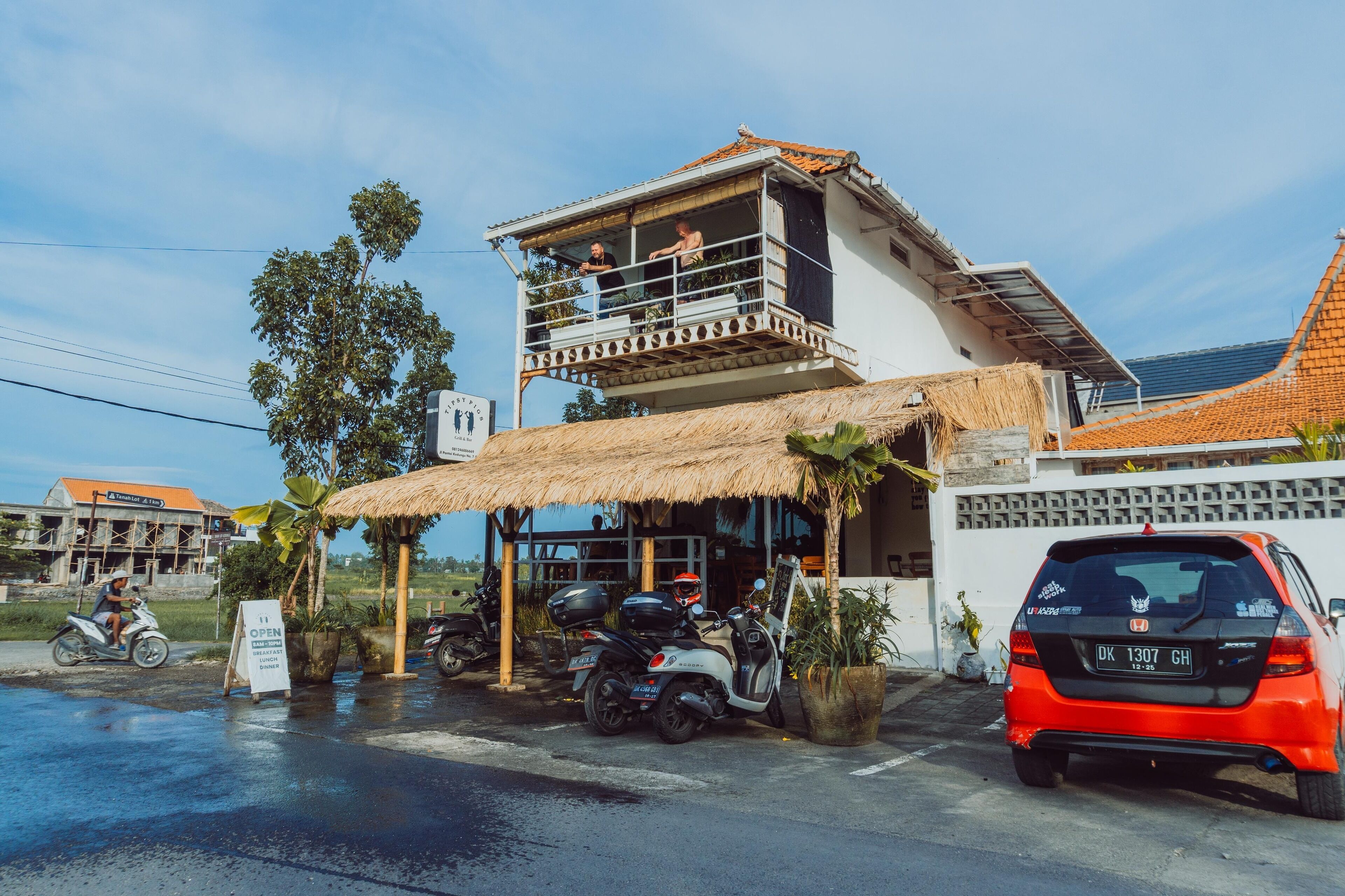 A building with a balcony, a thatched roof structure, and parked vehicles.