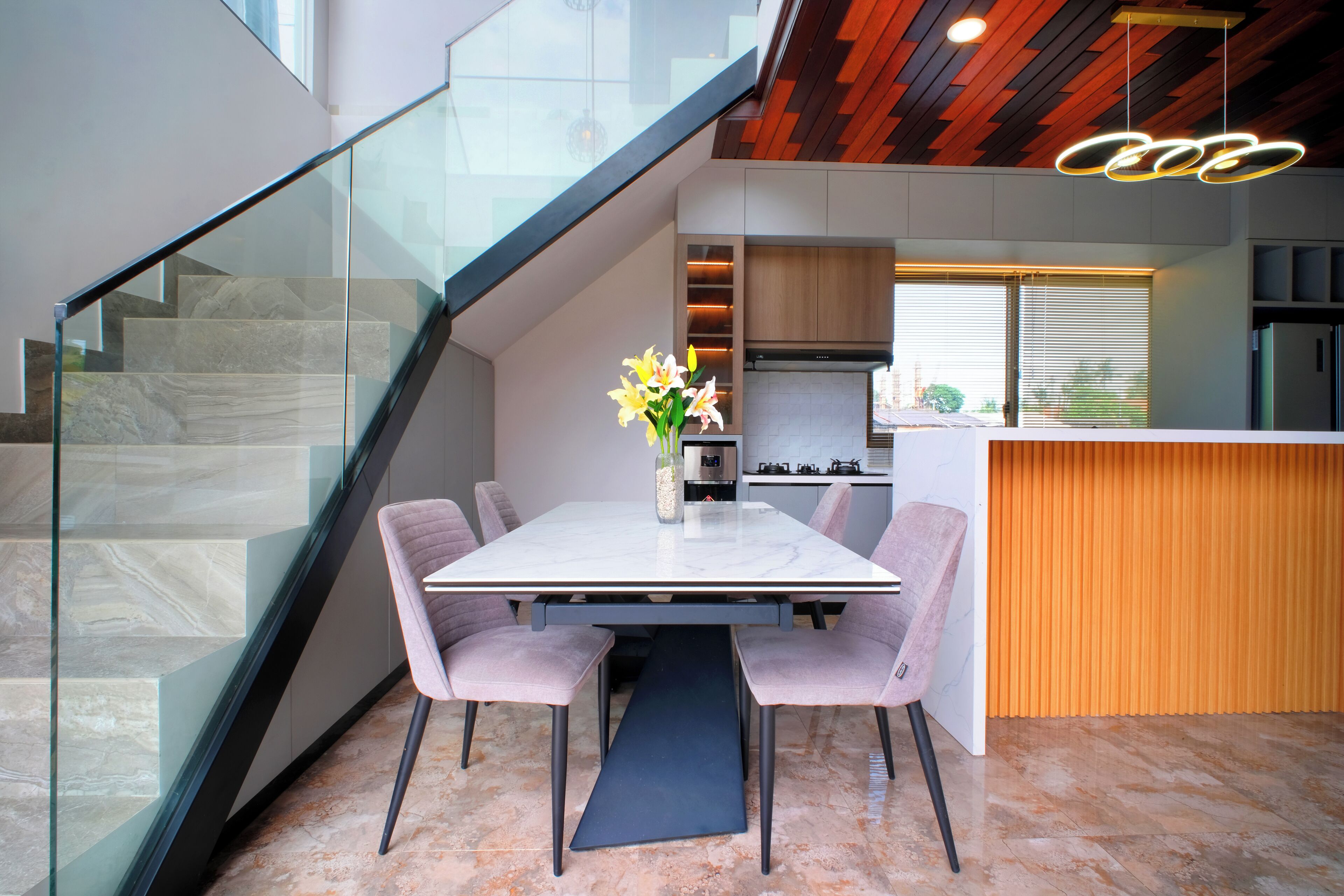 Une cuisine moderne dotée d’un îlot central, d’un coin repas avec une table en marbre et des chaises rembourrées, et d’un escalier avec des rampes en verre.