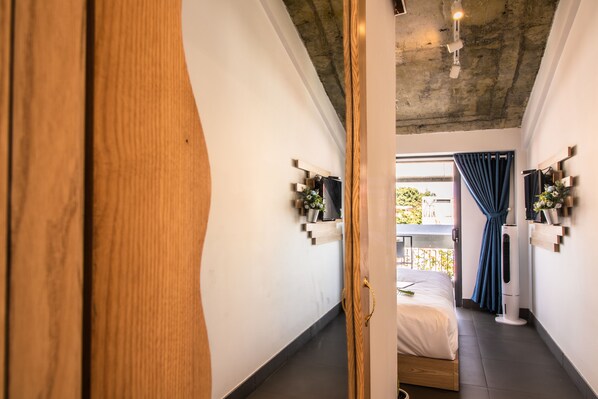 Corredor estreito que dá para um quarto, com cama, mesa de cabeceira e janela com cortinas.