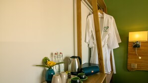 Habitación de hotel con escritorio de madera, espejo, bata blanca, lámpara, cafetera, tazas y un florero con flores.