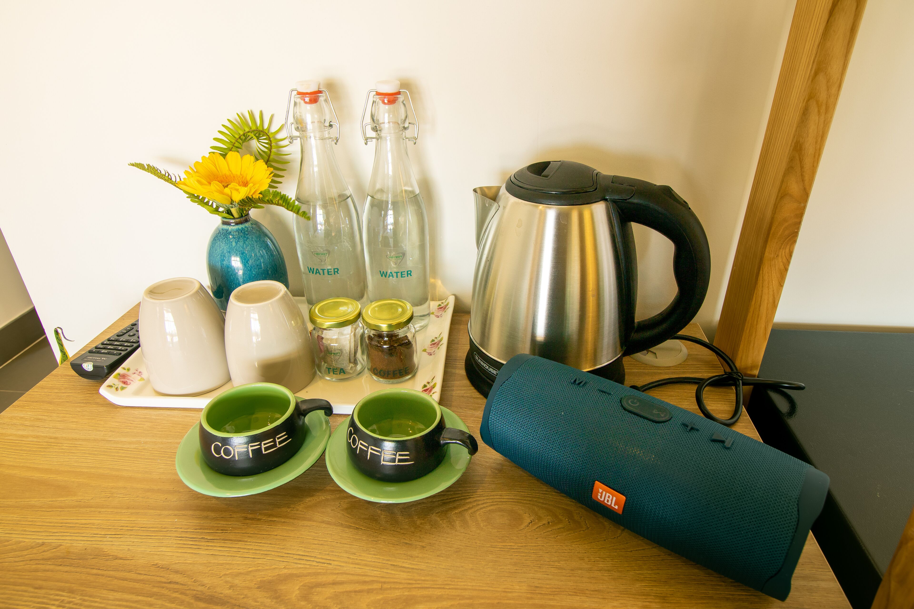 A wooden table with a coffee maker, two bottles of water, a vase with a yellow flower, and two mugs labeled "coffee".