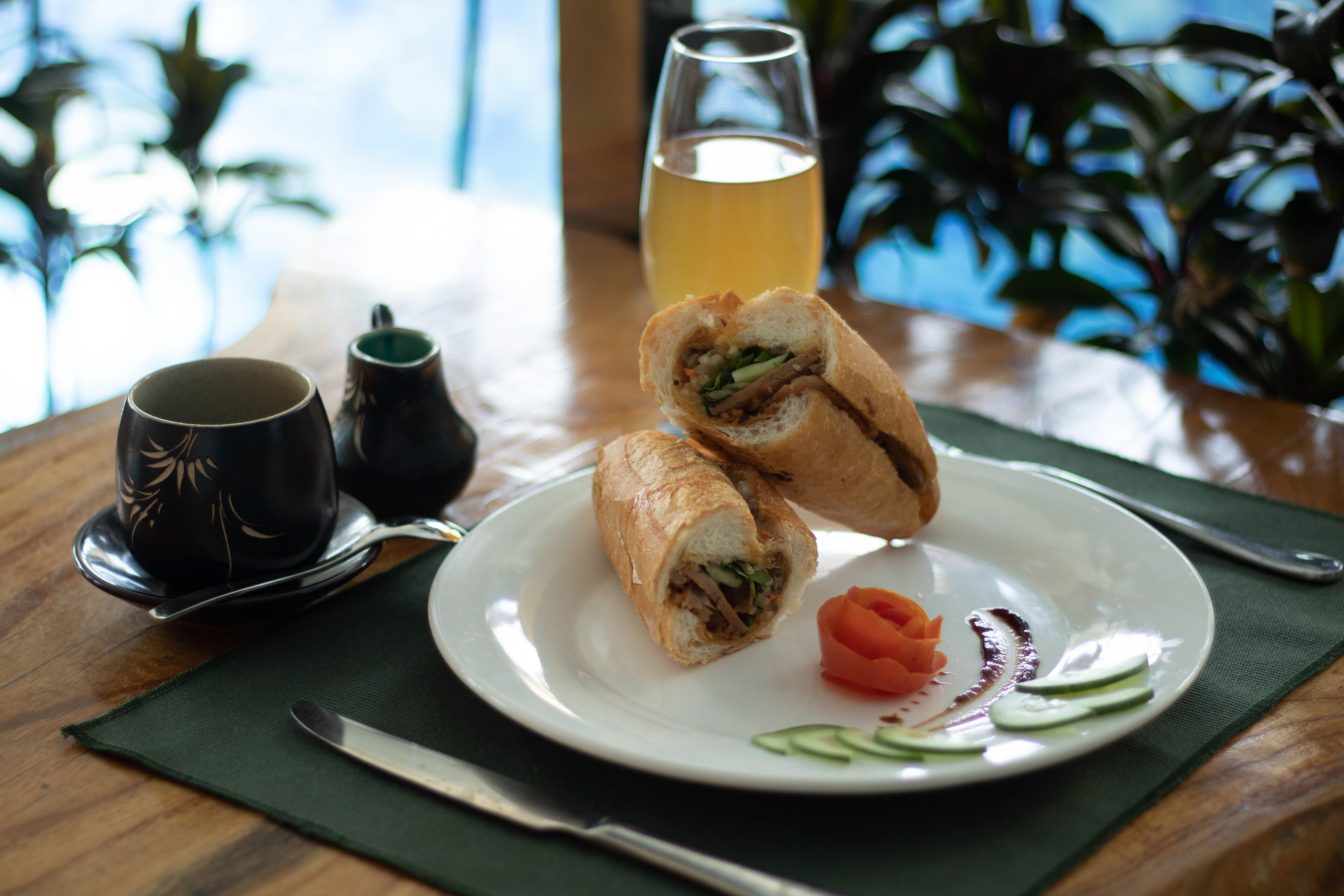 A table with a sandwich cut in half, a glass of yellow beverage, a cup, and a plate with some vegetables.