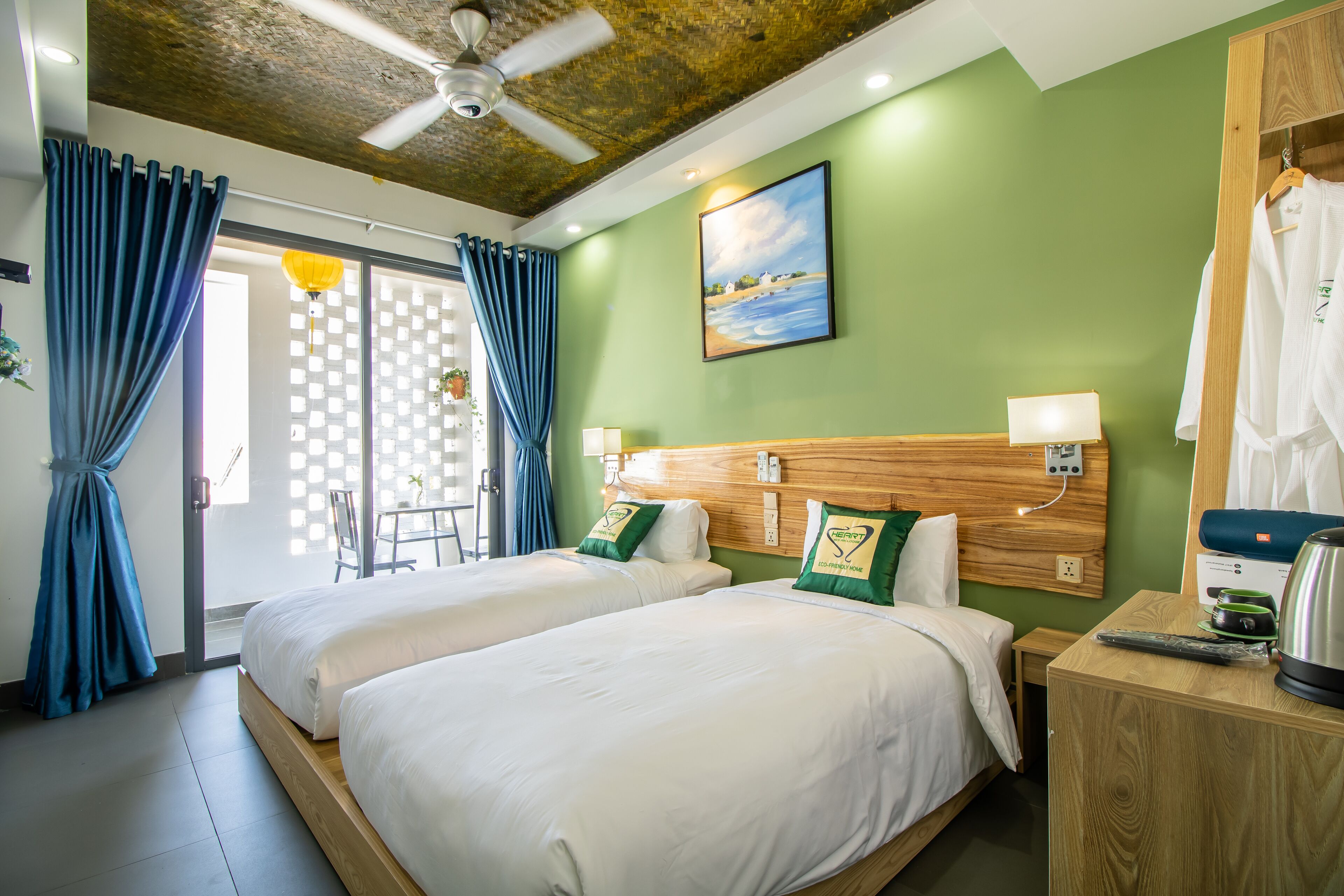 A hotel room with two beds, a wooden headboard, a painting, a ceiling fan, and a wooden nightstand with a kettle.