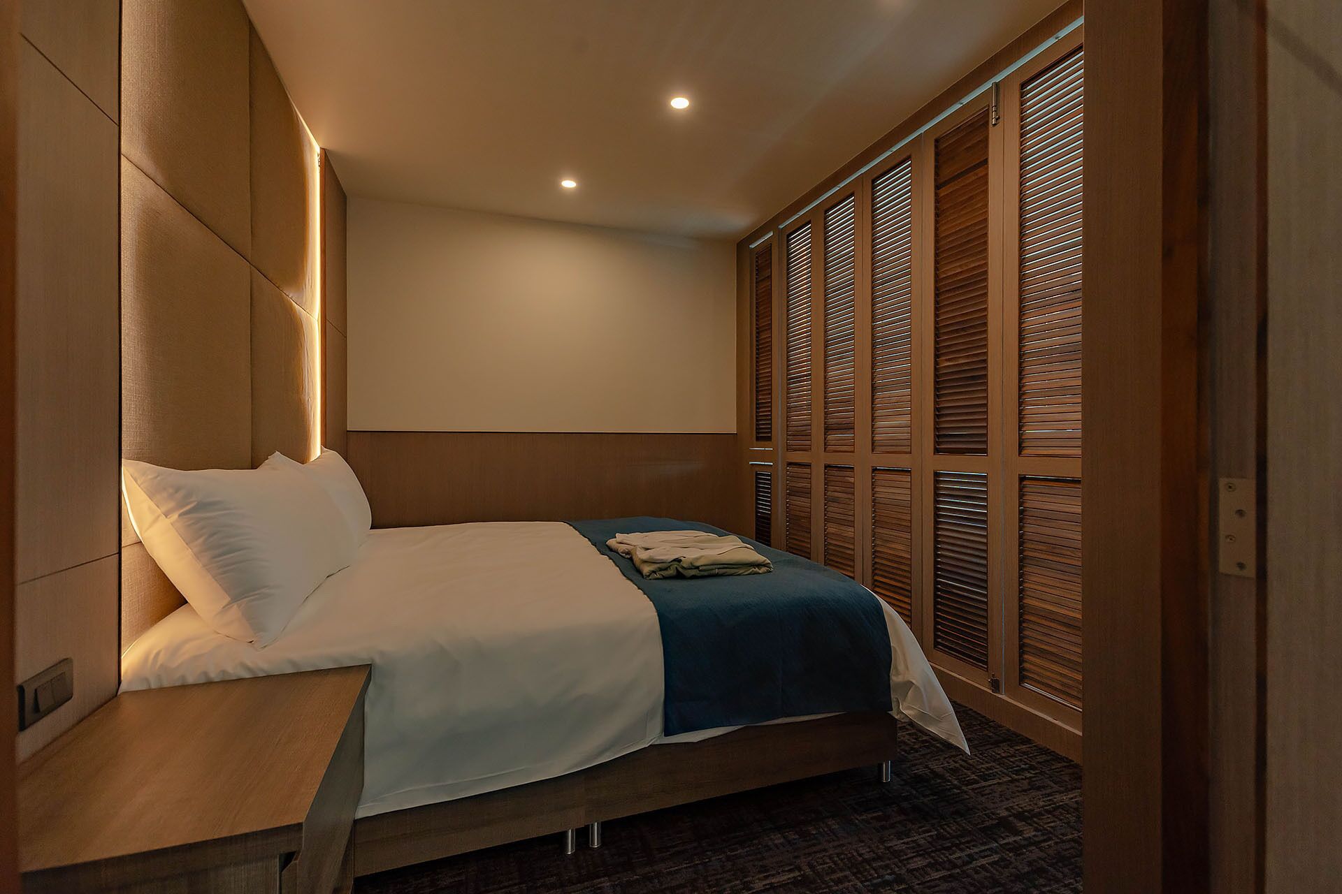 A hotel room with a bed, wooden headboard, and a large window with blinds.