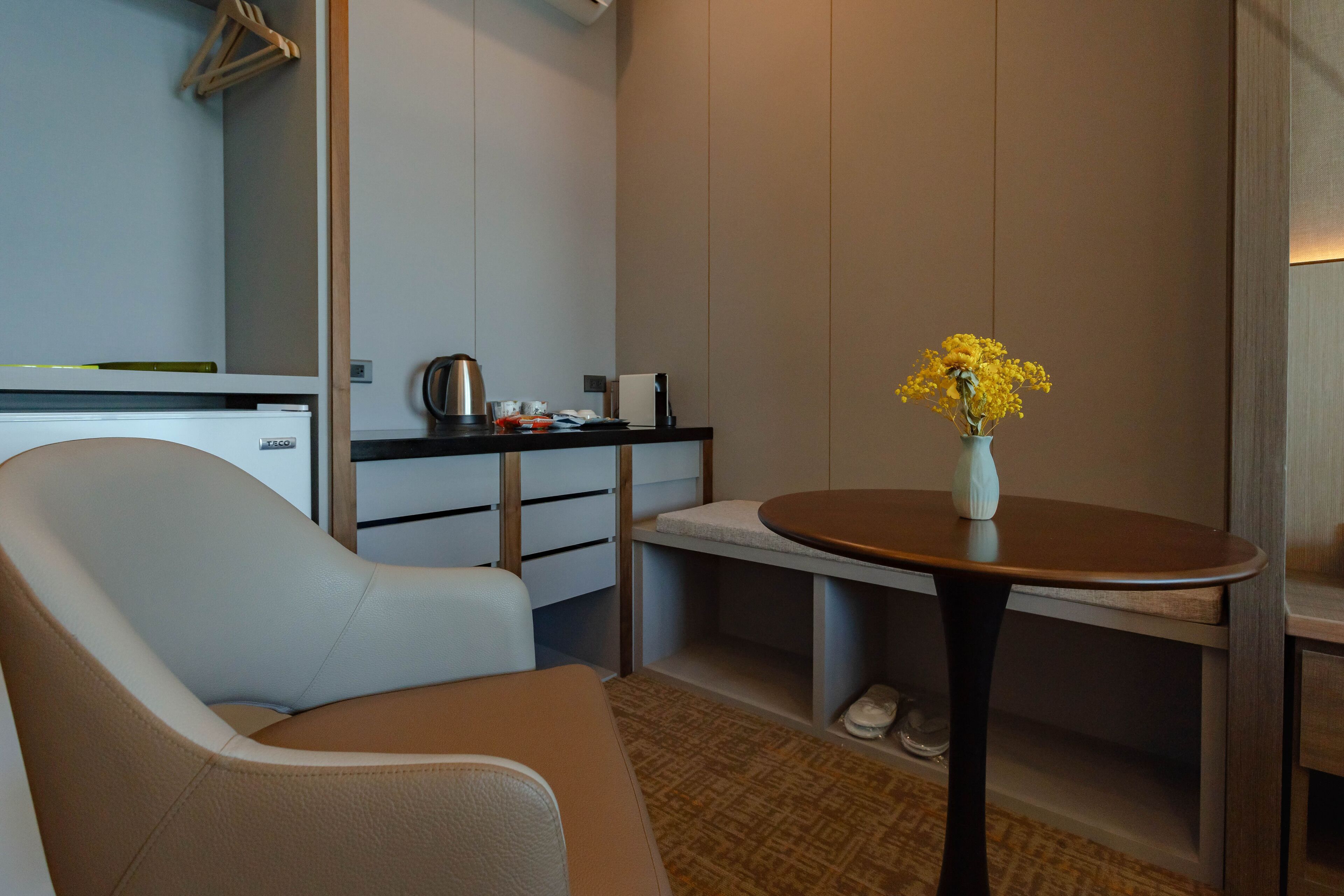 A modern hotel room with a sofa, a small round table with a vase of yellow flowers, a kitchenette, and a shoe rack.