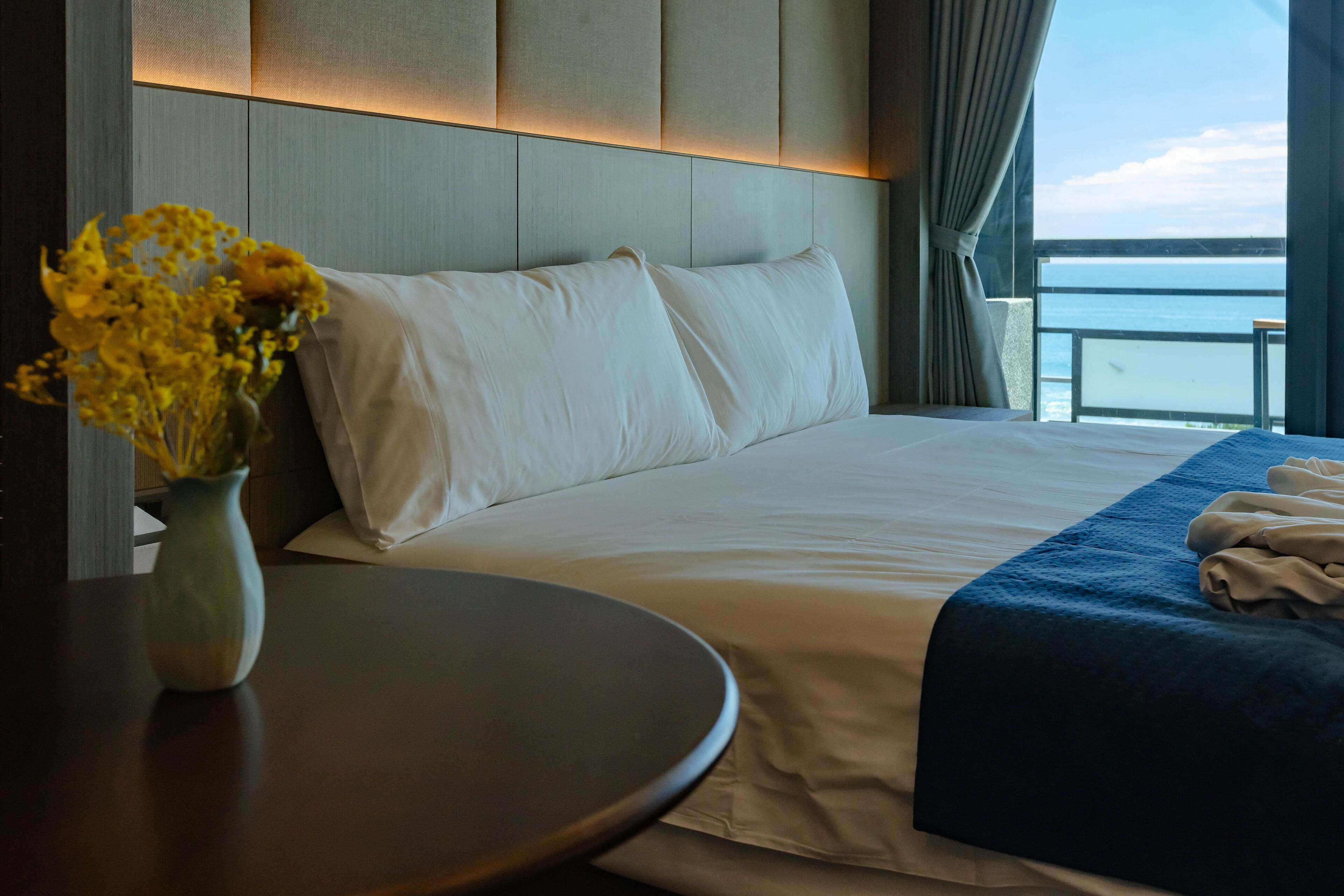 A hotel room with a large bed, a vase of yellow flowers, and a view of the sea.