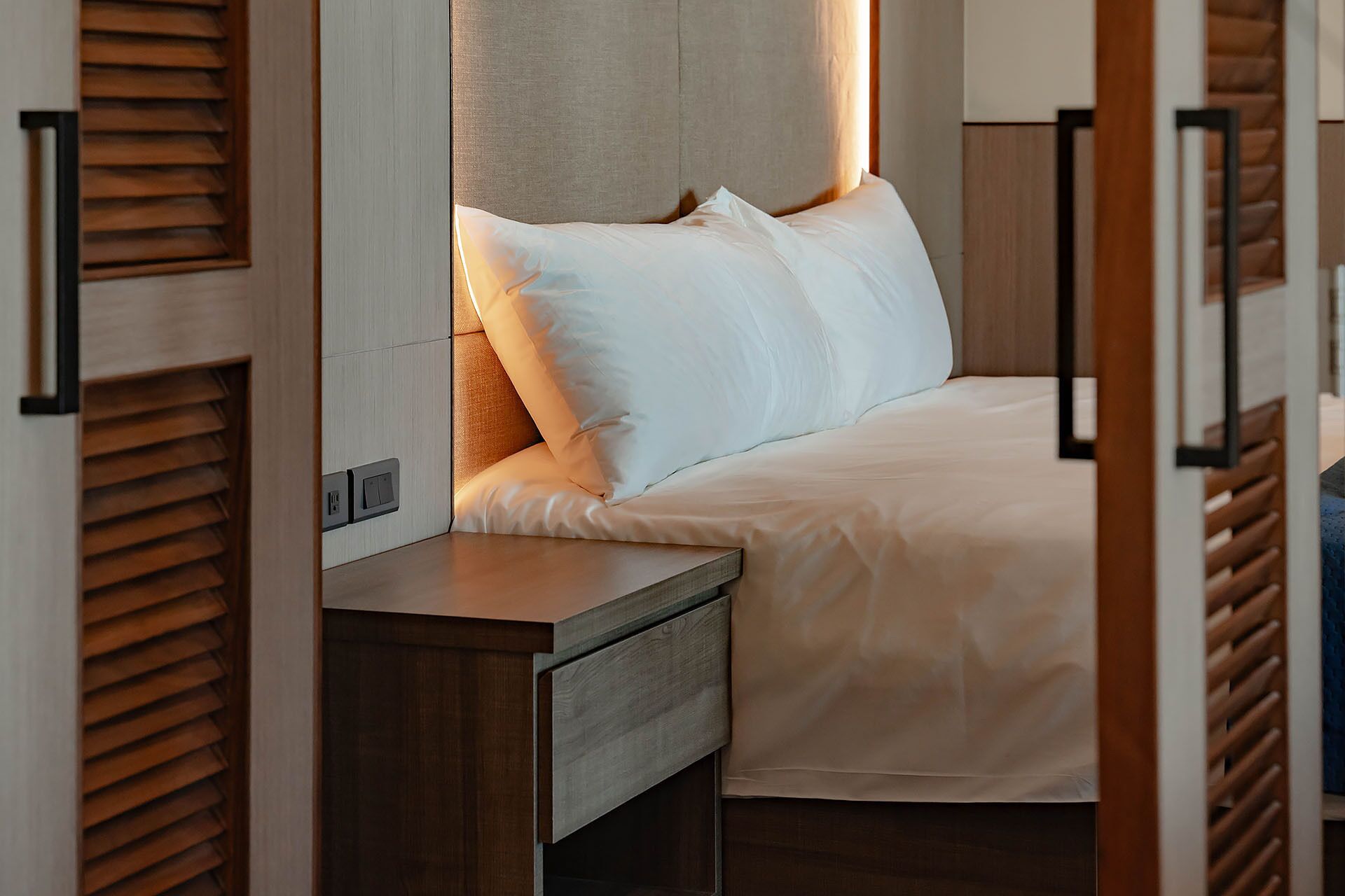 A neatly made bed with white linens, a wooden headboard, and a bedside table with a drawer.