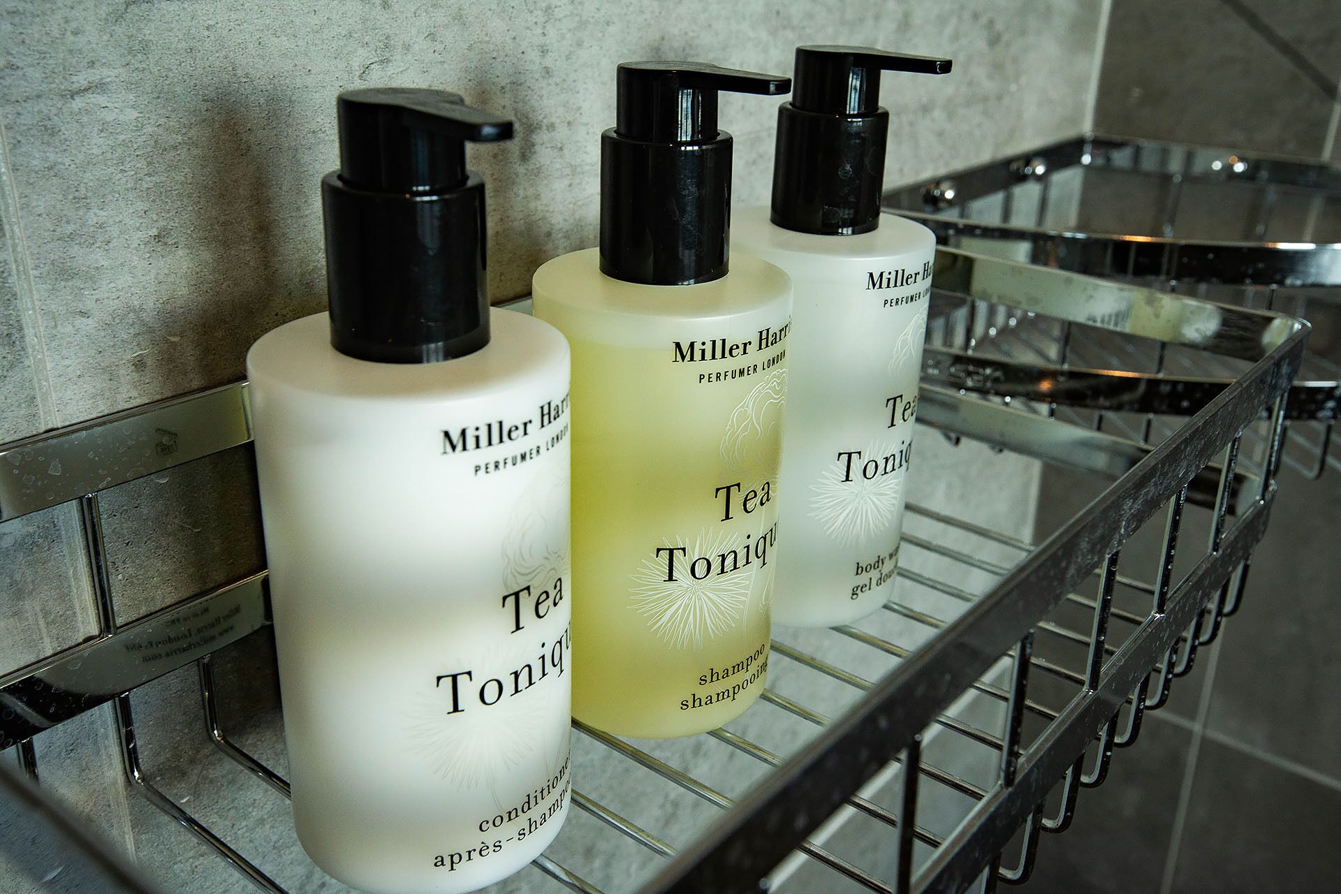 Trois flacons de shampooing Tea Tonique de la marque Miller Harris sont posés sur une étagère en métal dans une salle de bain.