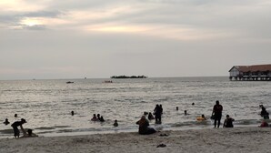 Ein Strand mit badenden und spielenden Personen im Wasser. In der Ferne ein Boot und ein Gebäude mit rotem Dach auf der rechten Seite.