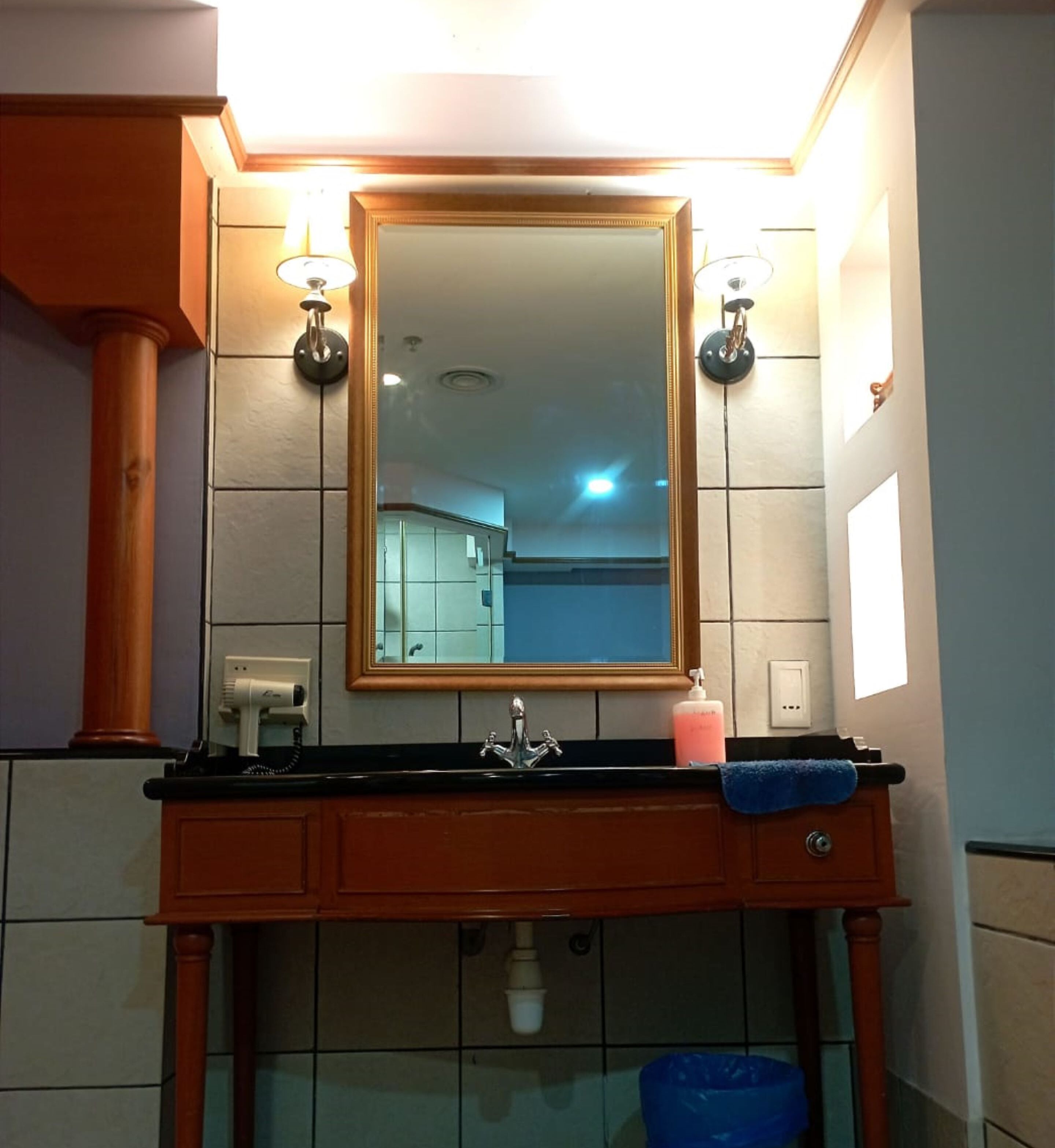 A bathroom with a wooden vanity, a mirror, two wall-mounted lights, a sink, a soap dispenser, and a blue towel.
