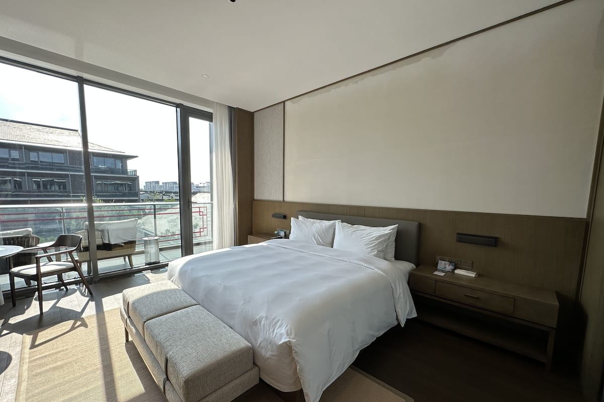 Una habitación de hotel moderna con una cama grande, una mesita de noche, un sillón y una zona de estar con un banco, y un balcón con vistas a la ciudad.