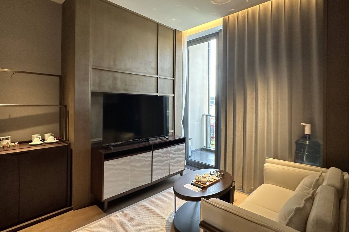 Habitación de hotel moderna con un televisor de pantalla plana grande, un sofá, una pequeña mesa redonda con un control remoto y vistas al exterior a través de un ventanal amplio.